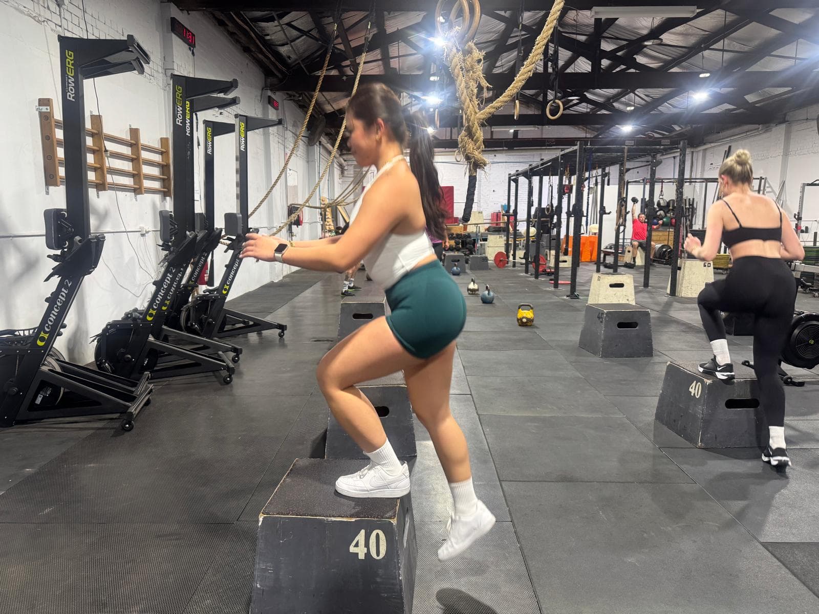 A member stepping onto a plyo box during a group class, rowing machines lining the wall
